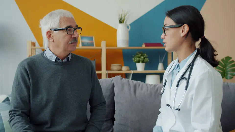 A clinician in a white coat talks with an older person on a sofa.