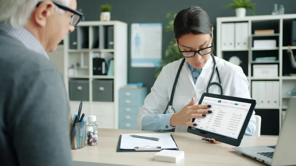 A clinician holds up a tablet showing a patient's results on the Cuneate dashboard while an older person looks on.
